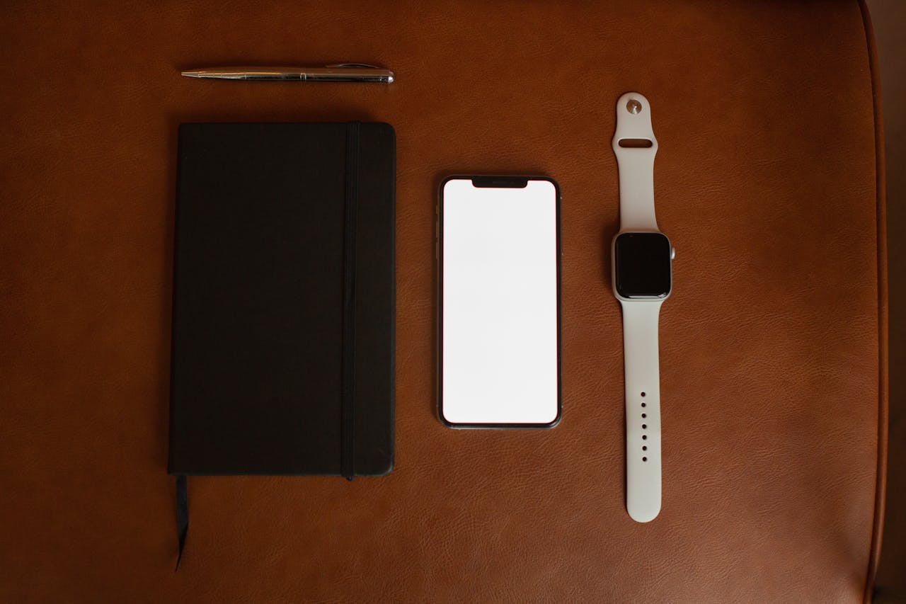 Flat lay of notebook, smartphone, and smartwatch on a leather surface for a modern workspace vibe.