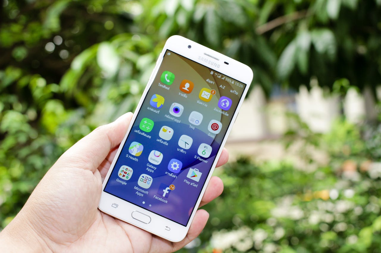Close-up of a hand holding a smartphone displaying various app icons against a blurred outdoor background.